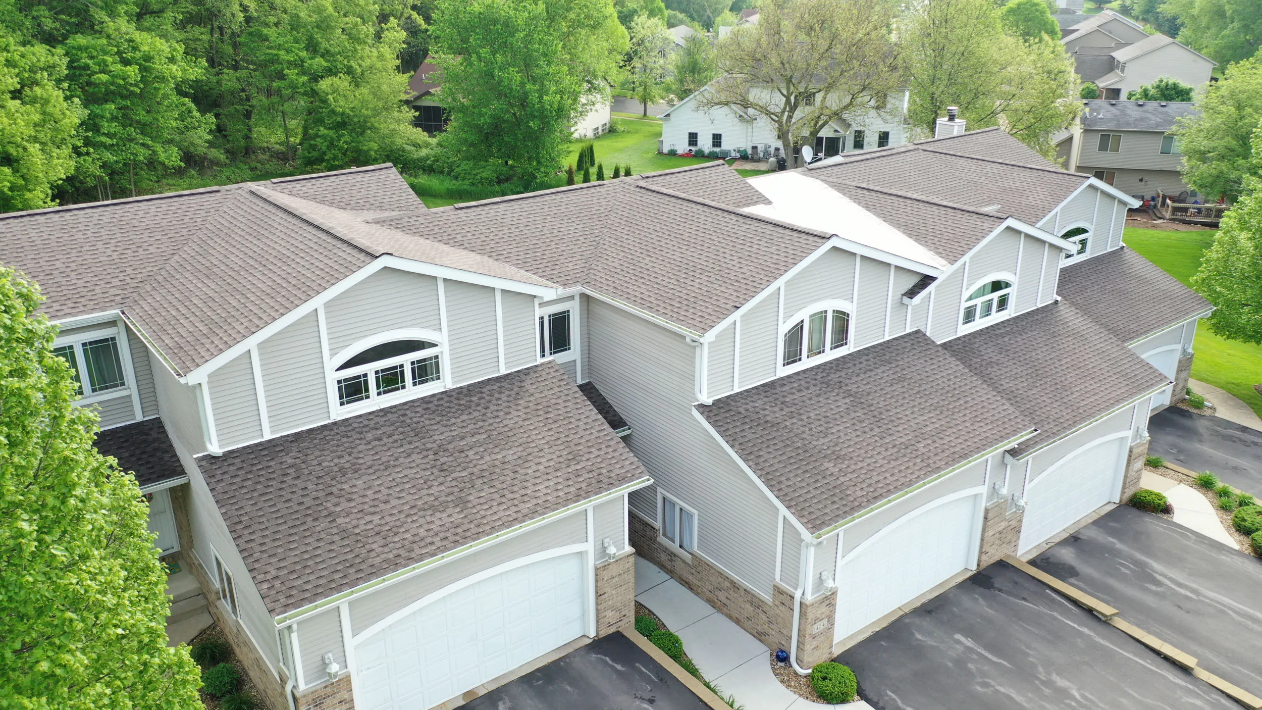 Aerial view of attached townhouses with gray roofs, white garage doors, and surrounding greenery.